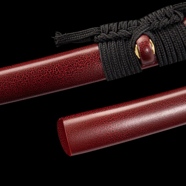 Peut inclure: Gros plan sur un fourreau de katana rouge avec une finition textur&eacute;e et craquel&eacute;e. Le fourreau est partiellement recouvert d'une gaine de cordon noir. Un accent de couleur or est visible pr&egrave;s de la poign&eacute;e. Le fond est noir.