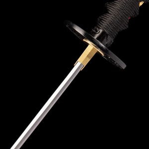 Peut inclure: Gros plan d'un katana japonais. La lame est argent&eacute;e, avec une garde en m&eacute;tal dor&eacute; et une poign&eacute;e noire envelopp&eacute;e de tissu noir. L'&eacute;p&eacute;e est pos&eacute;e sur un fond noir.