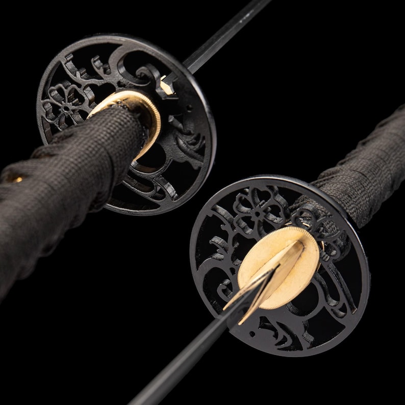 Peut inclure: Gros plan sur deux katanas japonais. Les &eacute;p&eacute;es ont des manches noirs envelopp&eacute;s de tissu noir, avec des gardes de main circulaires noires complexes. Les gardes de main pr&eacute;sentent des motifs floraux d&eacute;coup&eacute;s et des accents dor&eacute;s. Les lames sont noires et tranchantes.