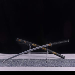 May include: A Japanese katana sword with a silver blade and black handle, resting in its black scabbard. The scabbard features gold dragon designs. The sword is displayed on a clear stand over a patterned cloth.