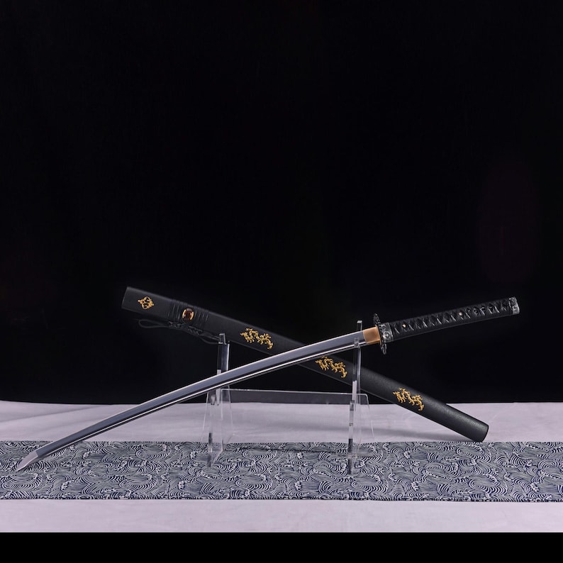 May include: A Japanese katana sword with a silver blade and black handle, resting in its black scabbard. The scabbard features gold dragon designs. The sword is displayed on a clear stand over a patterned cloth.