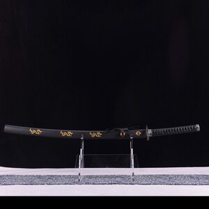 May include: A black katana sword with gold decorative accents, displayed on a clear stand. The sword's blade is curved, and the handle is wrapped with a textured material. The scabbard features gold floral designs.