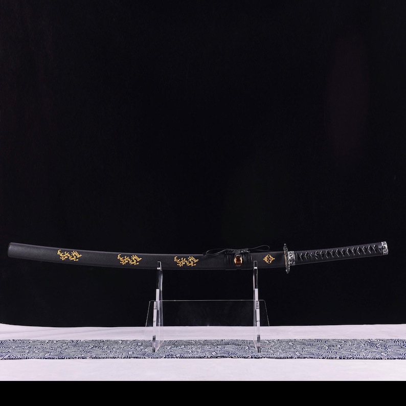 May include: A black katana sword with gold decorative accents, displayed on a clear stand. The sword's blade is curved, and the handle is wrapped with a textured material. The scabbard features gold floral designs.