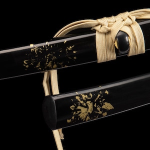 May include: Close-up of a black sword scabbard with gold floral accents. A gold-coloured cord is tied around the scabbard. The sword has a glossy finish against a black background.