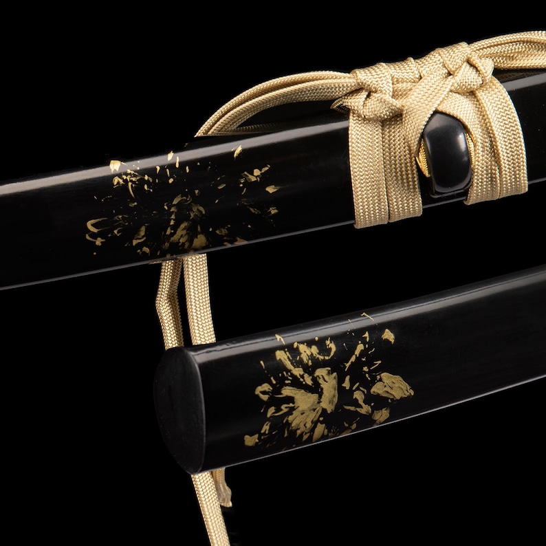 May include: Close-up of a black sword scabbard with gold floral accents. A gold-coloured cord is tied around the scabbard. The sword has a glossy finish against a black background.