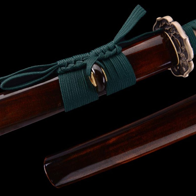 May include: A dark brown wooden katana scabbard with a green cord wrap and a decorative metal piece at the end.