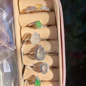 May include: A collection of gemstone rings displayed in a pink jewelry case. The rings feature various colored stones, including yellow, green, and pink, set in silver bands. Some rings are adorned with small, clear accent stones.