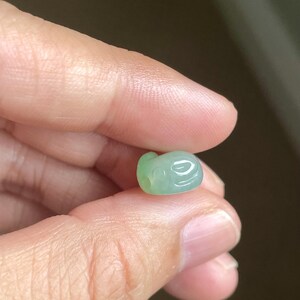 May include: A small, smooth, pale green carved bead with a translucent appearance. The bead features a rounded, organic shape with subtle indentations and a small circular hole visible on one side, showcasing a polished, glossy surface texture.