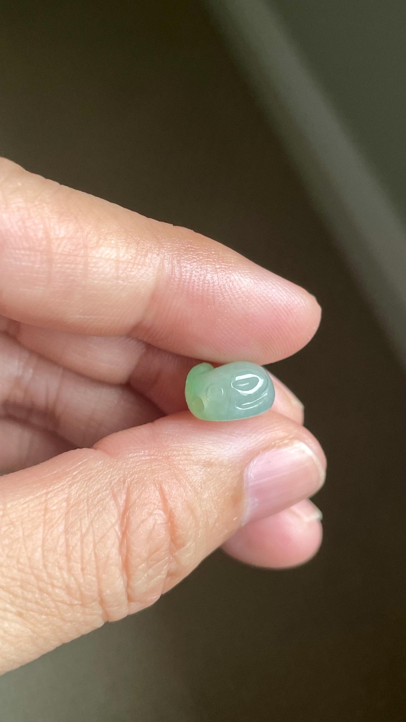 May include: A small, smooth, pale green carved bead with a translucent appearance. The bead features a rounded, organic shape with subtle indentations and a small circular hole visible on one side, showcasing a polished, glossy surface texture.