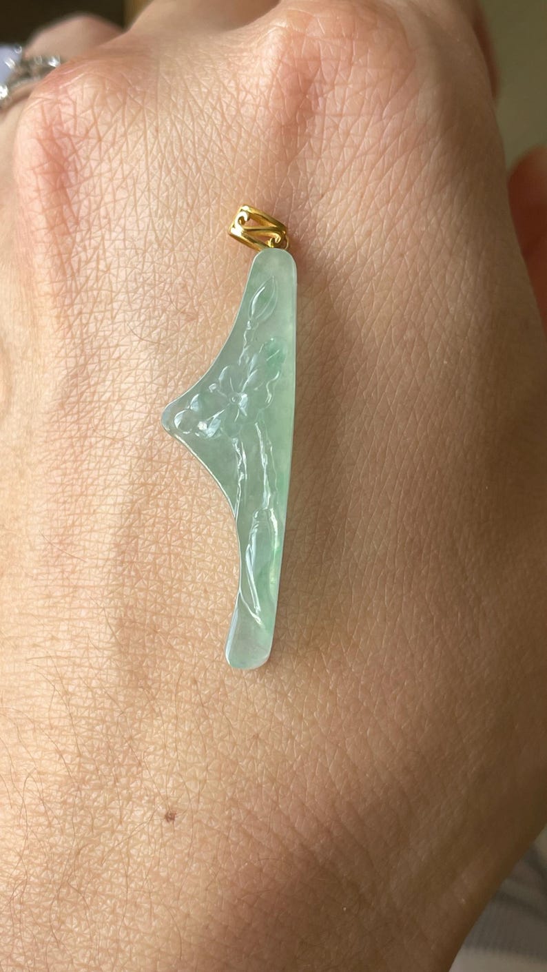 Peut inclure: Un pendentif en jade vert clair avec un motif floral. Le pendentif a une b&eacute;li&egrave;re dor&eacute;e en haut. Le jade est sculpt&eacute; d'un motif floral d&eacute;taill&eacute; et a une surface lisse et polie.