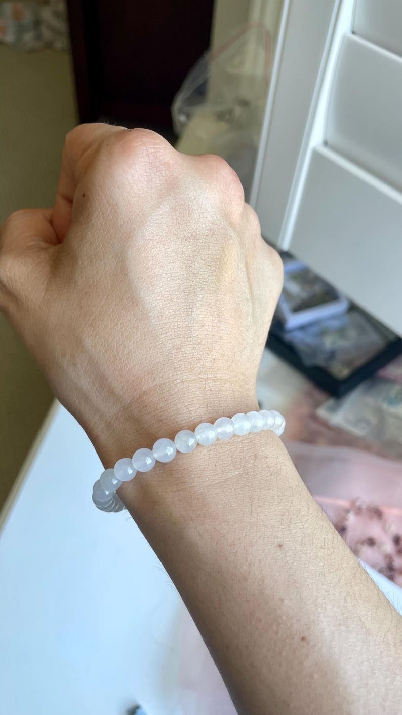 May include: A stretch bracelet composed of small, smooth, round, translucent white beads. The beads have a subtle, milky appearance and are strung together in a continuous circular loop, fitting snugly around the wrist.