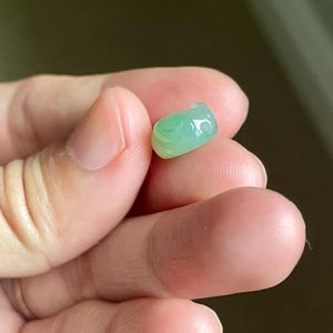 May include: A small, smooth, translucent light green jade bead with a polished surface and a rounded, cylindrical shape. The bead features subtle variations in color intensity, ranging from pale mint to a slightly deeper green hue.