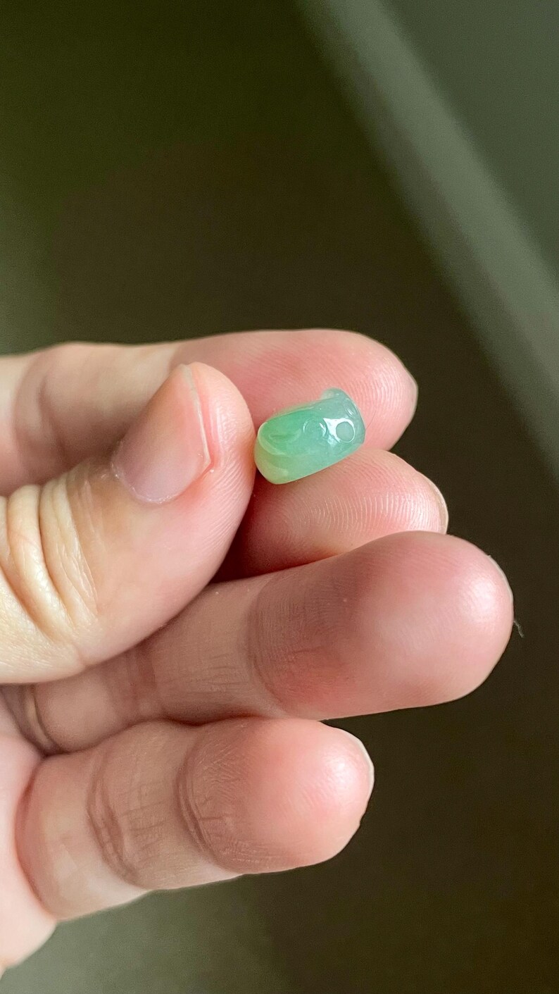 May include: A small, smooth, translucent light green jade bead with a polished surface and a rounded, cylindrical shape. The bead features subtle variations in color intensity, ranging from pale mint to a slightly deeper green hue.