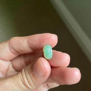 May include: A small, smooth, oval-shaped gemstone with a translucent, pale green color and a polished, glossy surface. The stone features subtle variations in light green tones throughout its rounded, bean-like form.