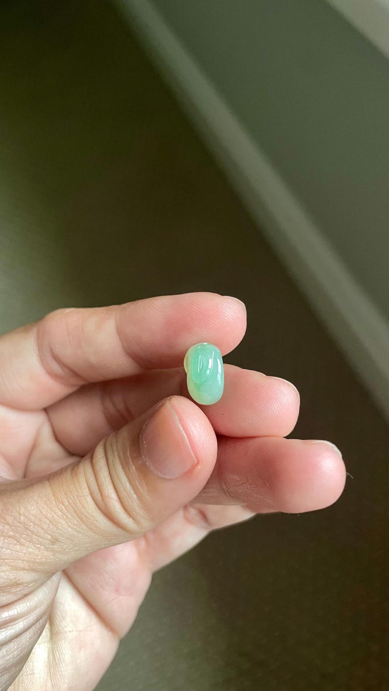 May include: A small, smooth, oval-shaped gemstone with a translucent, pale green color and a polished, glossy surface. The stone features subtle variations in light green tones throughout its rounded, bean-like form.