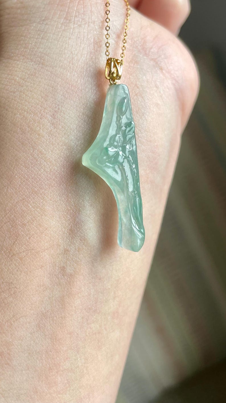 Peut inclure: Pendentif en jade vert clair avec un motif floral, suspendu &agrave; une fine cha&icirc;ne en or. Le pendentif a une surface lisse et polie. Le jade est d'une couleur vert p&acirc;le translucide. Le pendentif est pr&eacute;sent&eacute; sur un fond de peau.