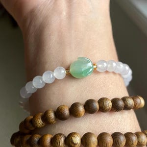 May include: A beaded bracelet featuring translucent white beads and a central green carved jade-like rabbit charm, paired with two stacked bracelets composed of round, dark brown wooden beads with a visible natural grain texture.