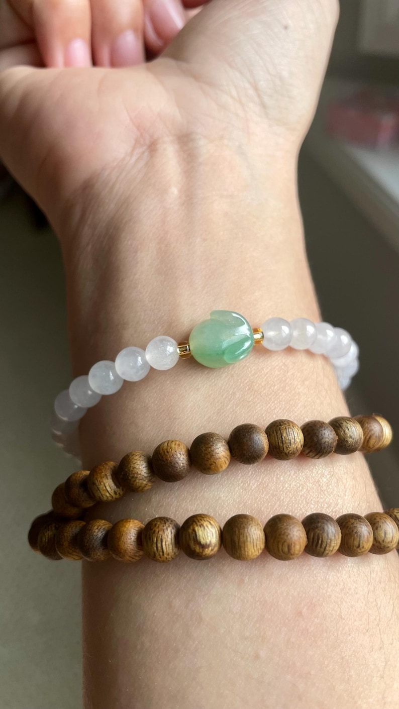 May include: A beaded bracelet featuring translucent white beads and a central green carved jade-like rabbit charm, paired with two stacked bracelets composed of round, dark brown wooden beads with a visible natural grain texture.
