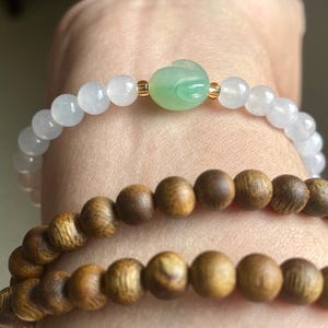 May include: A bracelet featuring translucent white beads and a central green carved jade rabbit bead with small gold-tone spacers, paired with a second bracelet made of round, dark brown, textured wooden beads.