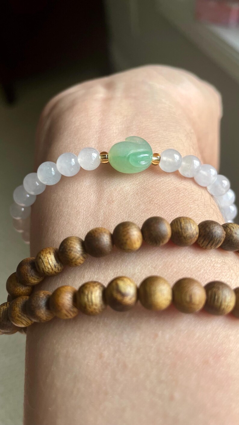 May include: A bracelet featuring translucent white beads and a central green carved jade rabbit bead with small gold-tone spacers, paired with a second bracelet made of round, dark brown, textured wooden beads.