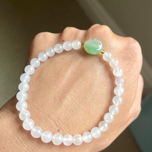 May include: A stretch bracelet composed of smooth, translucent white beads and a single carved green jade-style rabbit charm. The beads are strung on an elastic cord, creating a circular shape with a polished finish and a subtle, light-reflecting surface.