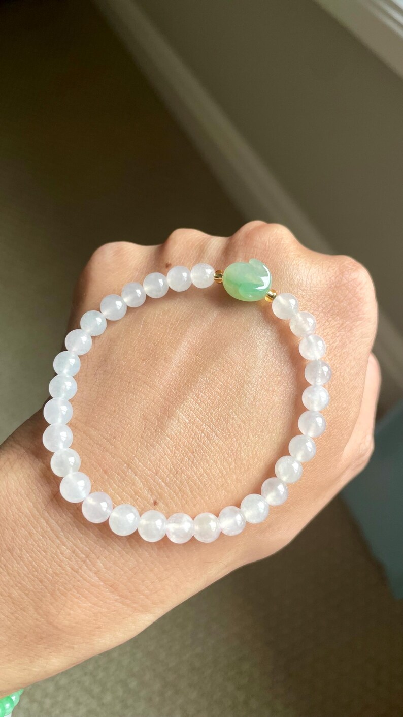 May include: A stretch bracelet composed of smooth, translucent white beads and a single carved green jade-style rabbit charm. The beads are strung on an elastic cord, creating a circular shape with a polished finish and a subtle, light-reflecting surface.