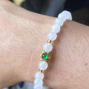 May include: A beaded bracelet featuring smooth, translucent white spherical beads and small gold-toned spacers. A central gold-toned star-shaped charm holds a small, round, vibrant green stone surrounded by tiny clear accent stones.