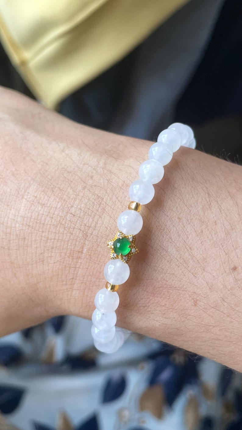 May include: A beaded bracelet featuring smooth, translucent white spherical beads and small gold-toned spacers. A central gold-toned star-shaped charm holds a small, round, vibrant green stone surrounded by tiny clear accent stones.