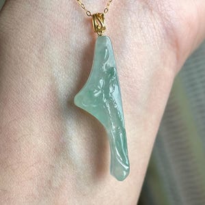 Peut inclure: Un collier en or d&eacute;licat avec un pendentif en jade vert clair. Le pendentif a une forme asym&eacute;trique unique avec des d&eacute;tails floraux sculpt&eacute;s. Le collier est pr&eacute;sent&eacute; sur un fond de ton chair, soulignant l'&eacute;l&eacute;gance du bijou.
