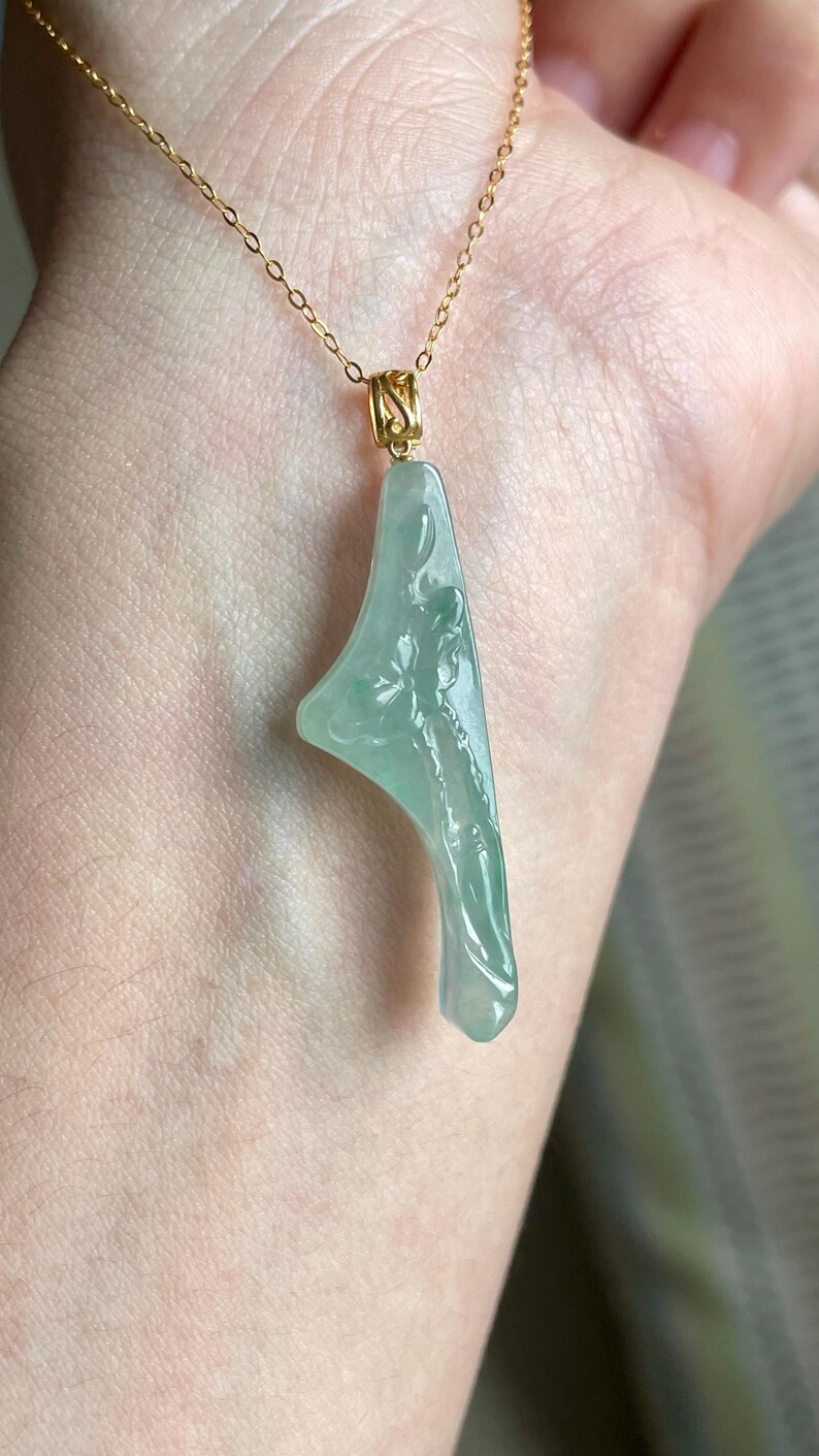 Peut inclure: Un collier en or d&eacute;licat avec un pendentif en jade vert clair. Le pendentif a une forme asym&eacute;trique unique avec des d&eacute;tails floraux sculpt&eacute;s. Le collier est pr&eacute;sent&eacute; sur un fond de ton chair, soulignant l'&eacute;l&eacute;gance du bijou.