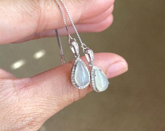 Peut inclure: Une paire de boucles d'oreilles pendantes argentées avec des pierres vert pâle en forme de goutte. Chaque pierre est entourée d'un halo de petits cristaux clairs et étincelants sertis dans une monture métallique, suspendue à une fine chaîne.