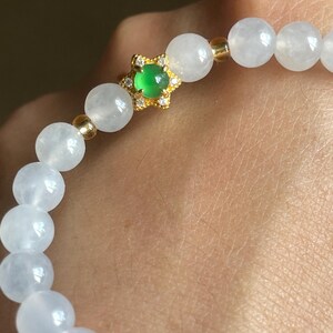 May include: A beaded bracelet featuring smooth, translucent white spherical beads interspersed with small gold-toned spacers. A central gold-toned star-shaped charm holds a small, vibrant green circular stone surrounded by tiny clear crystals.
