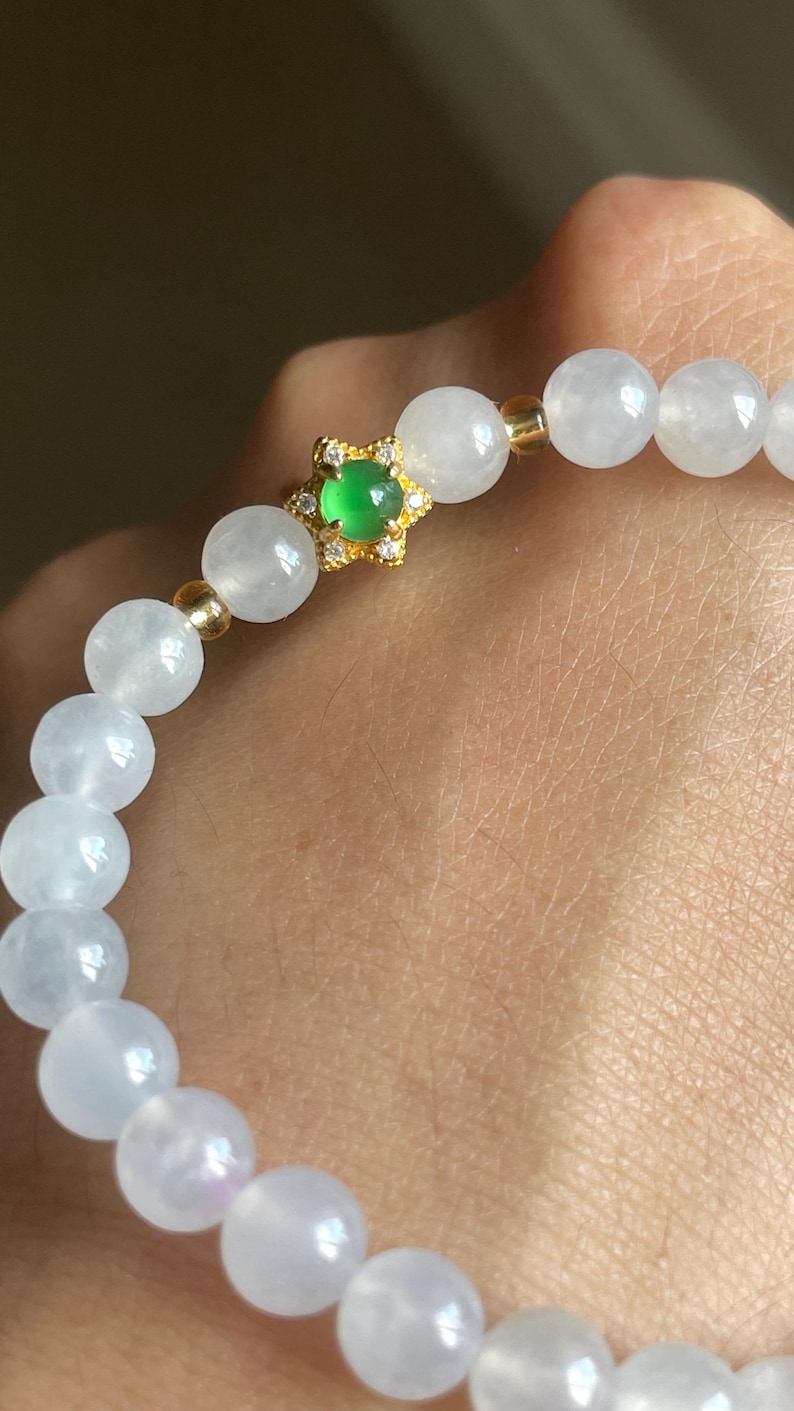 May include: A beaded bracelet featuring smooth, translucent white spherical beads interspersed with small gold-toned spacers. A central gold-toned star-shaped charm holds a small, vibrant green circular stone surrounded by tiny clear crystals.