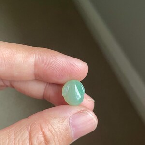 May include: A small, smooth, light green jade gemstone carved into the shape of a rabbit head with two elongated ears pointing downwards. The stone has a polished, translucent finish and a rounded, oval-like silhouette.