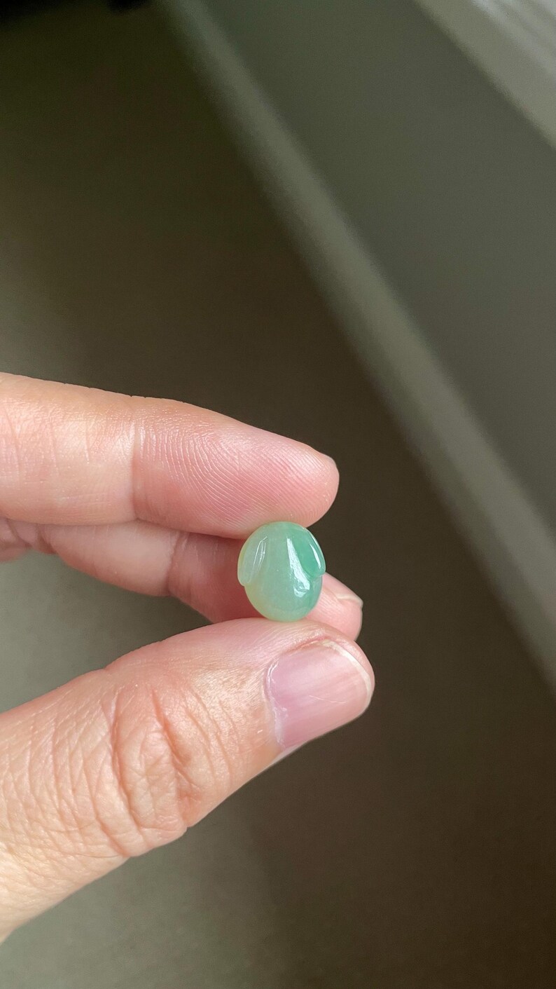 May include: A small, smooth, light green jade gemstone carved into the shape of a rabbit head with two elongated ears pointing downwards. The stone has a polished, translucent finish and a rounded, oval-like silhouette.