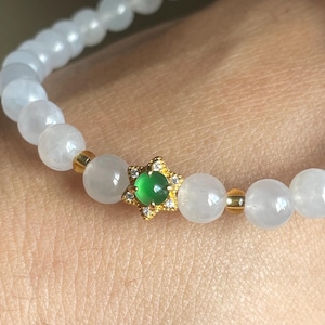 May include: A beaded bracelet featuring smooth, translucent white spherical beads interspersed with small gold-toned spacers. A central gold-toned star-shaped charm holds a small, round, vibrant green cabochon stone accented with tiny clear crystals.