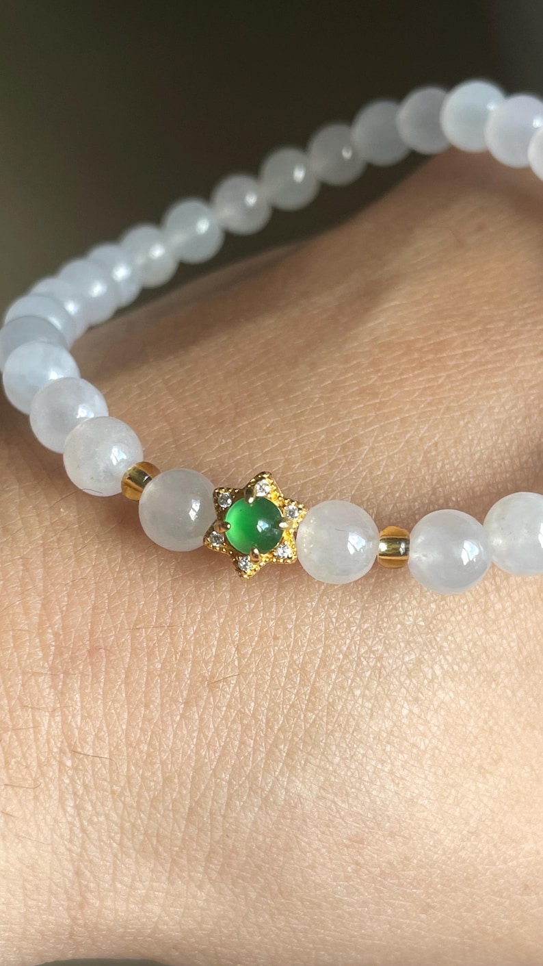 May include: A beaded bracelet featuring smooth, translucent white spherical beads interspersed with small gold-toned spacers. A central gold-toned star-shaped charm holds a small, round, vibrant green cabochon stone accented with tiny clear crystals.