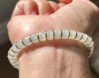 May include: A bracelet composed of alternating smooth, translucent white, square-shaped beads and thin, polished gold-colored metal spacers. The beads have a soft, milky appearance and are arranged in a continuous circular pattern around the wrist.