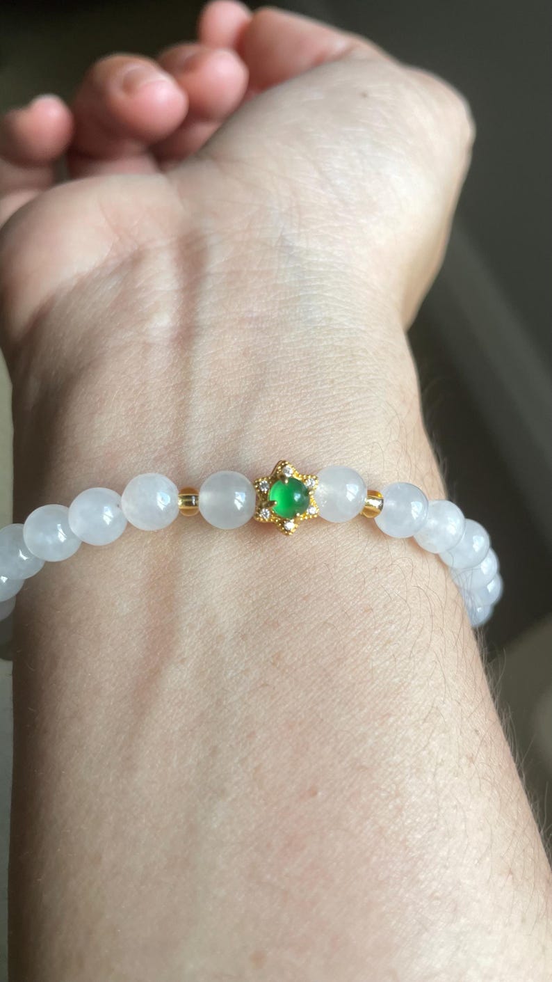 May include: A beaded bracelet featuring smooth, translucent white round beads and a central gold-toned star-shaped charm. The charm is set with a small, vibrant green cabochon stone at its center, surrounded by tiny clear accent stones.