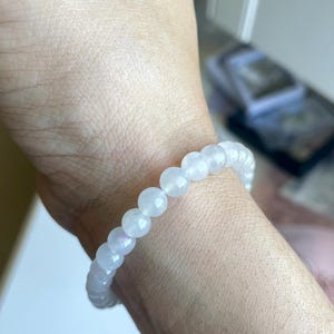 May include: A stretch bracelet composed of smooth, round, translucent white beads with a subtle iridescent sheen. The beads are uniform in size and strung together in a continuous circular loop, resting against the skin of a wrist.