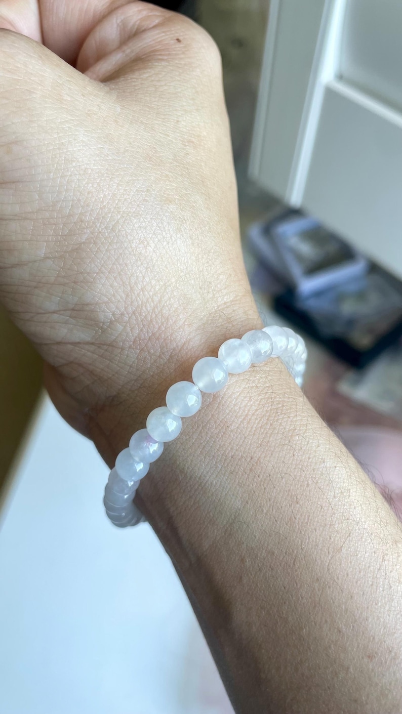 May include: A stretch bracelet composed of smooth, round, translucent white beads with a subtle iridescent sheen. The beads are uniform in size and strung together in a continuous circular loop, resting against the skin of a wrist.