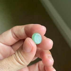 May include: A small, smooth, oval-shaped cabochon made of translucent light green stone. The piece features a polished, rounded surface with subtle variations in color intensity, displaying a soft, pale mint-green hue throughout its compact form.