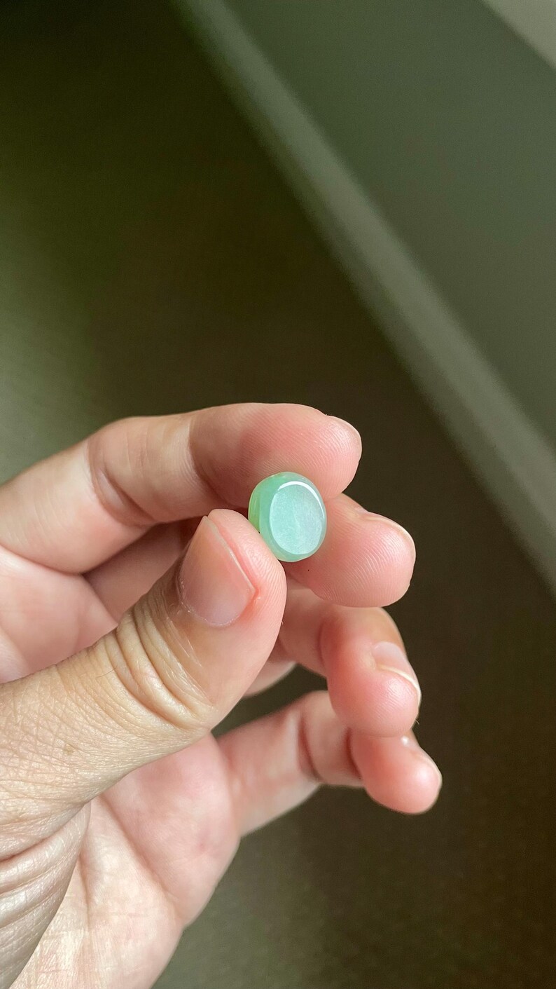 May include: A small, smooth, oval-shaped cabochon made of translucent light green stone. The piece features a polished, rounded surface with subtle variations in color intensity, displaying a soft, pale mint-green hue throughout its compact form.