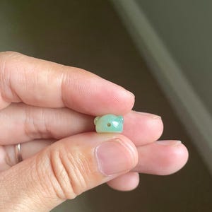 May include: A small, smooth, light green jade bead carved into the shape of a bear head. The bead features two rounded ears and a single horizontal hole drilled through the center of the face for stringing purposes.