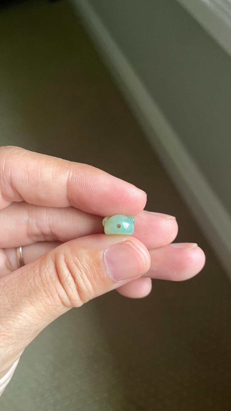May include: A small, smooth, light green jade bead carved into the shape of a bear head. The bead features two rounded ears and a single horizontal hole drilled through the center of the face for stringing purposes.