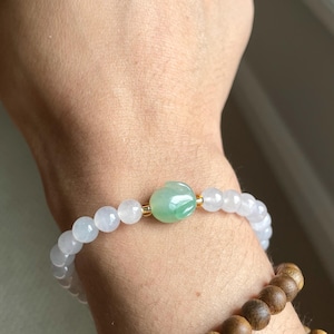 May include: A beaded bracelet featuring translucent pale lavender round beads and a central carved green jade-like bead with gold-toned spacers. Below it, a second bracelet consists of dark brown wooden beads with a natural grain texture.