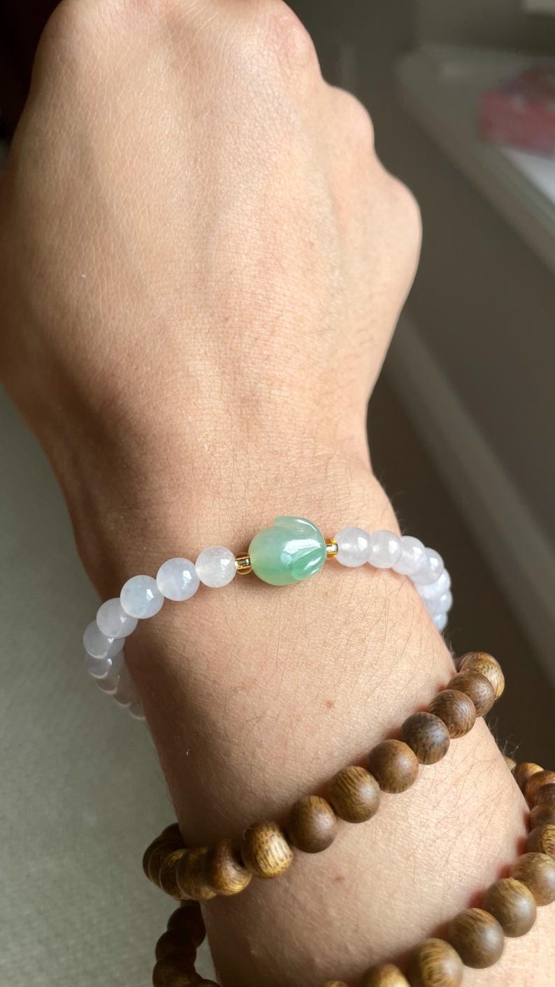 May include: A beaded bracelet featuring translucent pale lavender round beads and a central carved green jade-like bead with gold-toned spacers. Below it, a second bracelet consists of dark brown wooden beads with a natural grain texture.