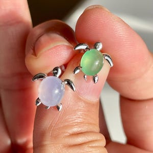 May include: A pair of turtle-shaped stud earrings. One earring features a light green stone, the other a light purple stone. The turtle's body is a smooth, rounded stone, and the legs and head are silver-colored metal.