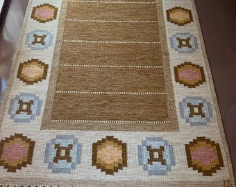 May include: A rectangular woven rug with a central brown field and a patterned border. The border features repeating geometric shapes in shades of pink, blue, and brown against a cream background. The rug has a fringe along the shorter edges.