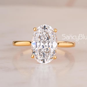 May include: A gold ring featuring a large, clear, oval-cut center stone held by four prongs on a thin, polished gold band. The text SancyBlue is visible to the right of the ring, which is centered against a light, neutral background.